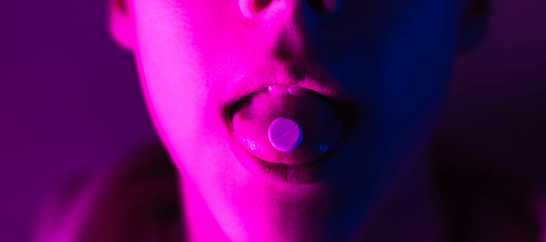 Person mit einer 1D-LSD-Tablette auf der Zunge, geöffneter Mund, vor einem Hintergrund in lebhaften, bunten Farben, was die psychedelische Erfahrung symbolisiert.